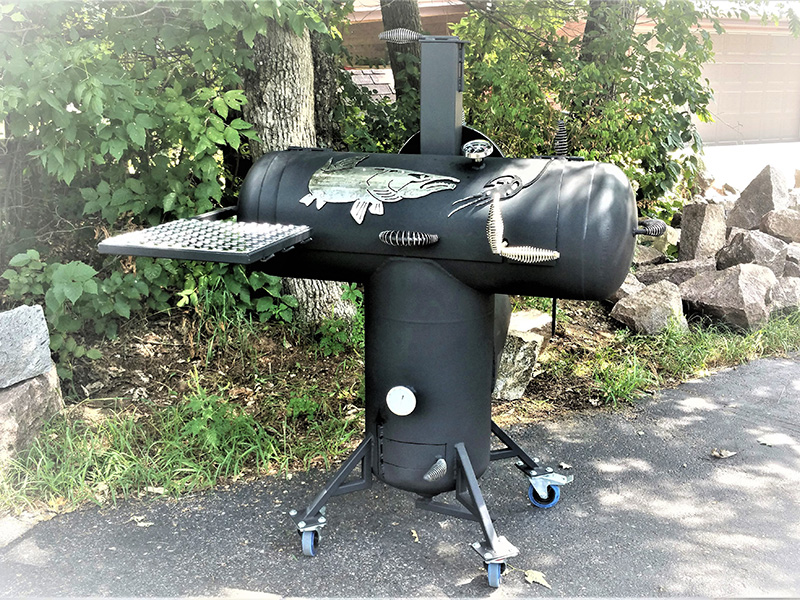 T-shaped charcoal-color grill w/ silver-color cover art center muskie & lure. Background trees w/ green leaves, rocks, and paved driveway.