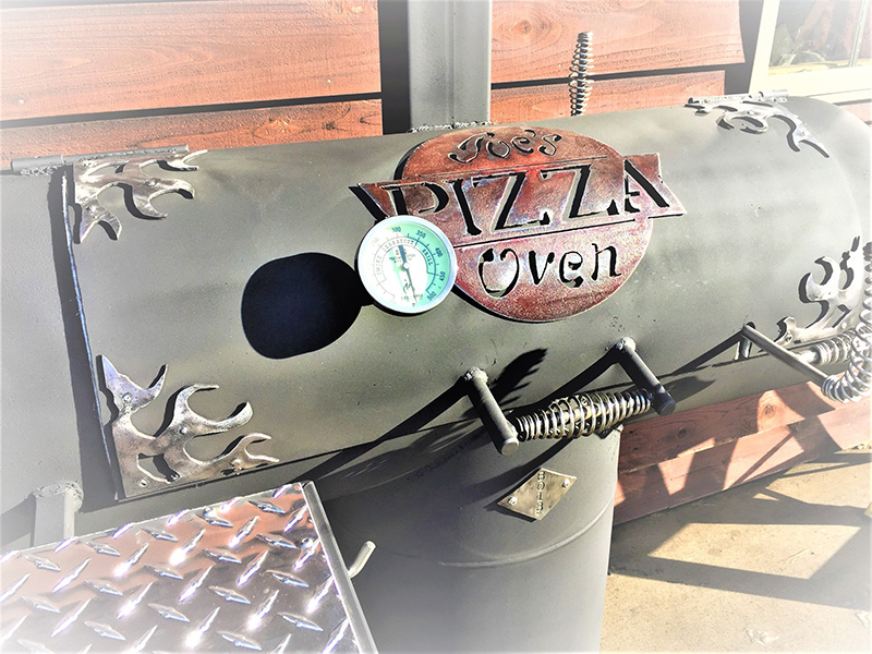 T-shaped charcoal-color grill w/ cover art flames in corners, center red plaque Pizza Oven. Background horisontal red cedar exterior siding.