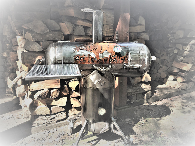 T-shaped gunmetal-color grill w/ rust-color cover art boar & words Get In My Belly. Background woodshed with stacked split wood.