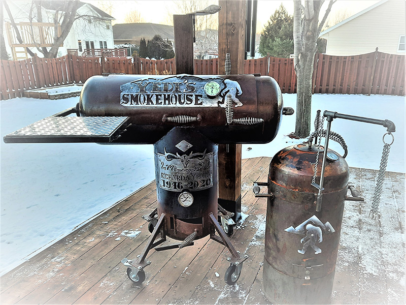 T-shaped rust-color grill w/ silver front shelf and cover art that says Yetis Smokehouse. Next to a rust colored Bullet grill