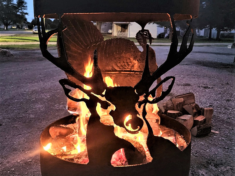 Round black firepit with flames in center. Front art cutout stag. Back art cutout turkey